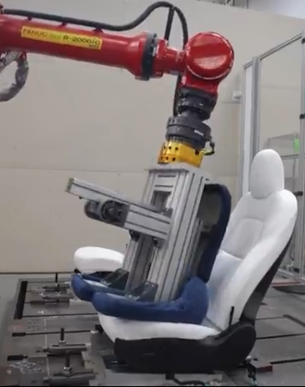 FANUC R-2000iC robot arm with aluminum extrusion torso-shaped end effector performing ingress/egress durability testing on a Tesla seat clamped to a T-slot fixture table